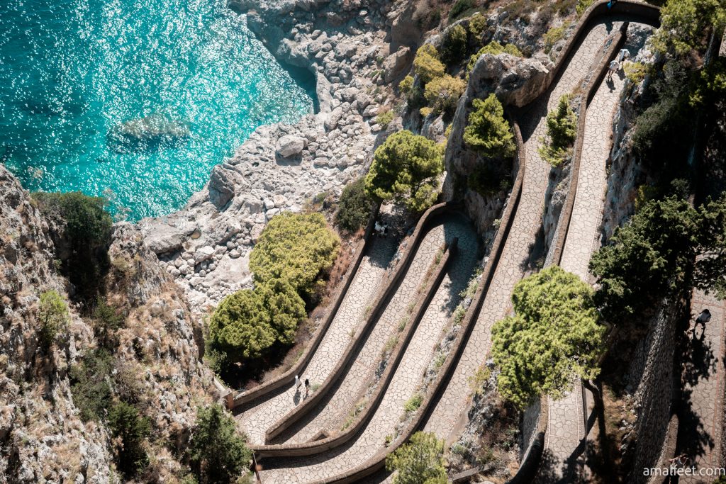 A Via Krupp felülnézeből. egymást érő hajtűkanyarok meredek sziklába vájva, mediterrán növényzet és türkiz kék tenger, sziklás parttal látható a képen.