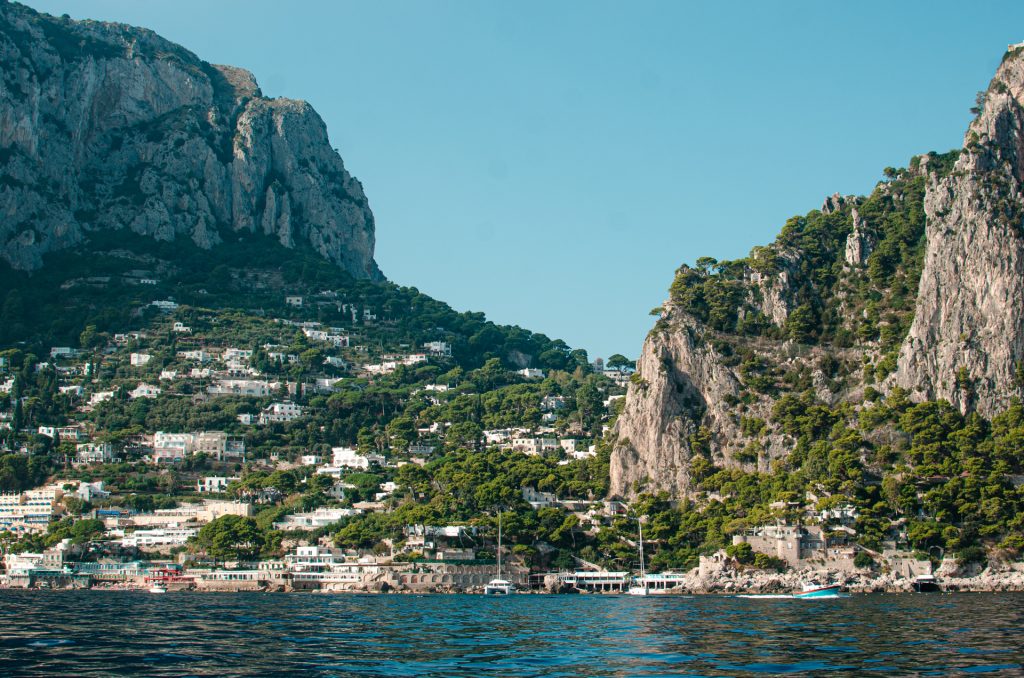 Capri, látképe a tengerről. Zöld erdős terület sok hófehér villával itt-ott elszórva a tájban. Hatalmas sziklafalak.