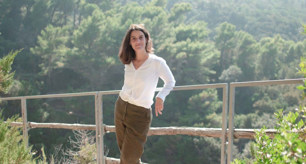 Én ahogy egy fa korláthoz támaszkodom egy ösvényen Capri szigetén. Fehér blúzt és khaki zöld len nadrágot viselek. A háttér kissé elmosódott, napfényes fenyőerdő
